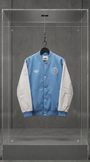 Jaqueta varsity Adidas x Inter Miami azul-clara com mangas creme, vista frontal, em vitrine de acrílico